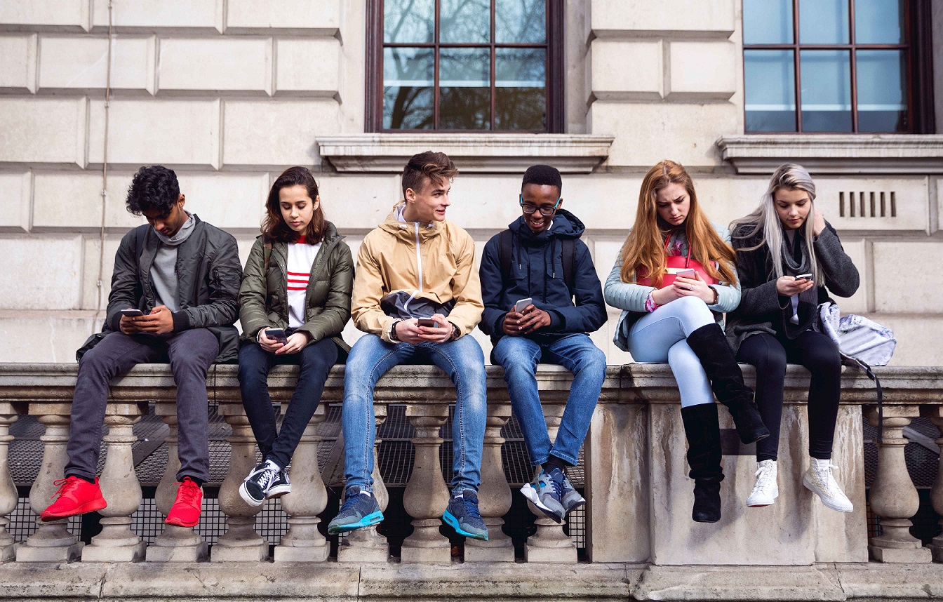 high-school-aged kids sitting along a building's stone rail, looking at mobile devices